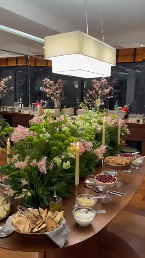 Mesa de buffet à noite sob um lustre, com arranjos verdes e rosa, velas e tortas de romã